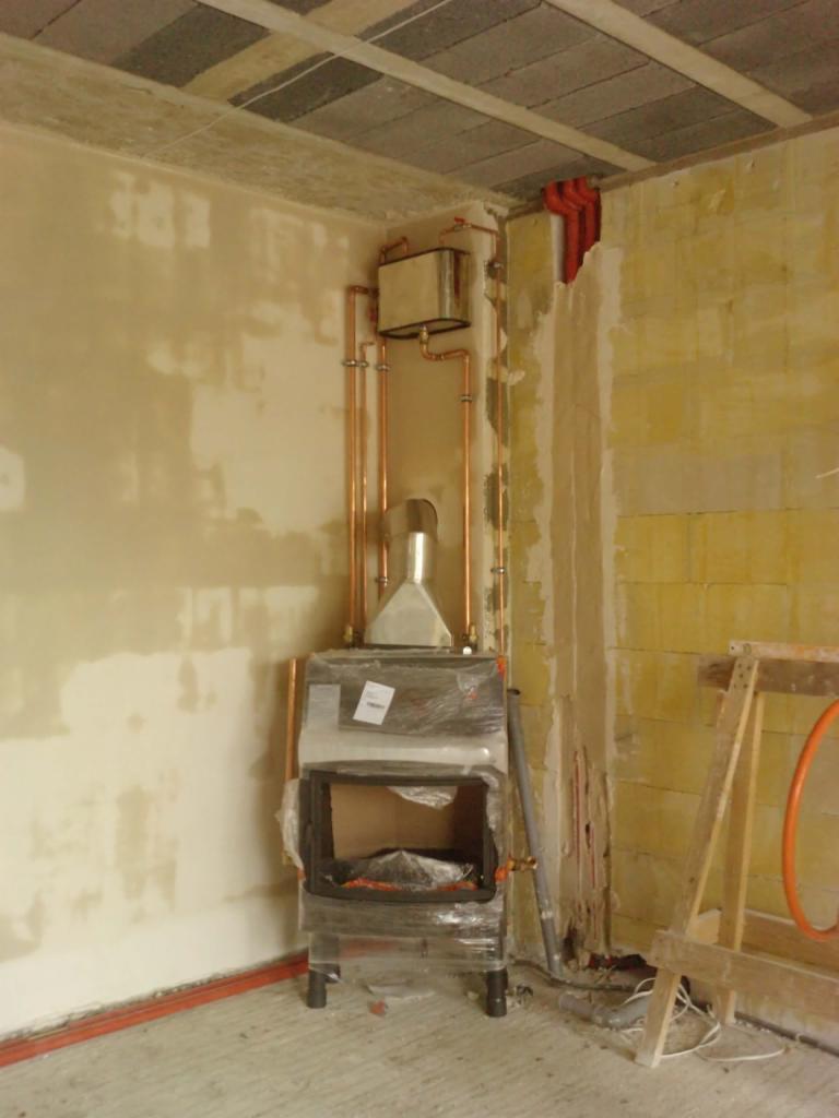 Nowy piec kominkowy w folii ochronnej, z podłączonymi miedzianymi rurami instalacji grzewczej w surowym wnętrzu, w trakcie prac wykończeniowych.