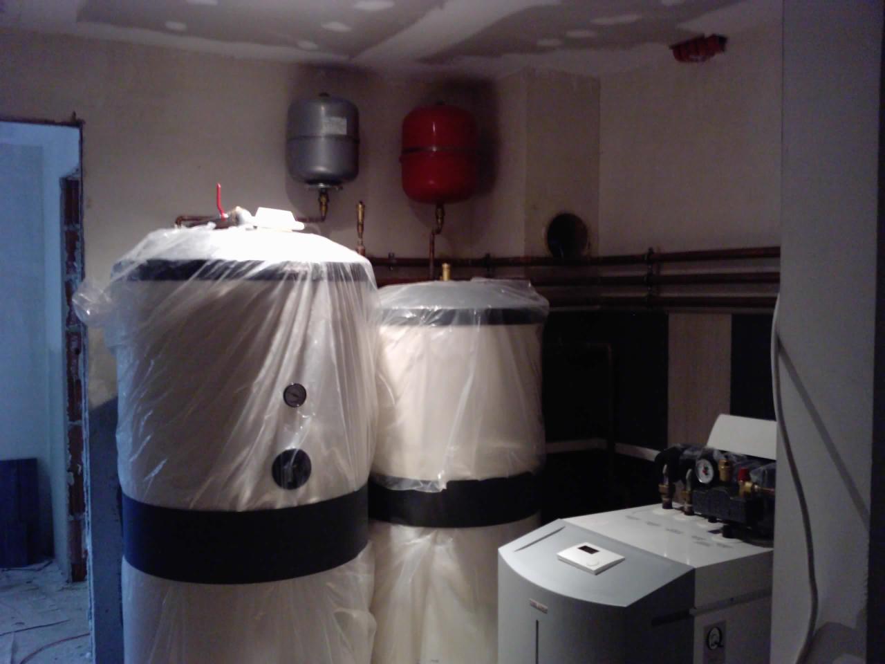 Dwa białe zbiorniki z czarnymi pasami w folii ochronnej, rury miedziane na ścianie, szary piec grzewczy. Elementy instalacji grzewczej w surowym pomieszczeniu.