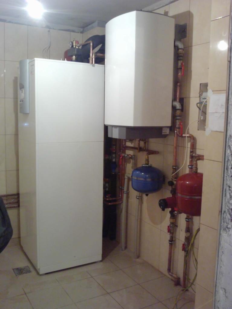 Nowoczesna instalacja grzewcza z białym zbiornikiem i piecem, z widocznymi miedzianymi rurami oraz elementami armatury w pomieszczeniu wyłożonym płytkami.
