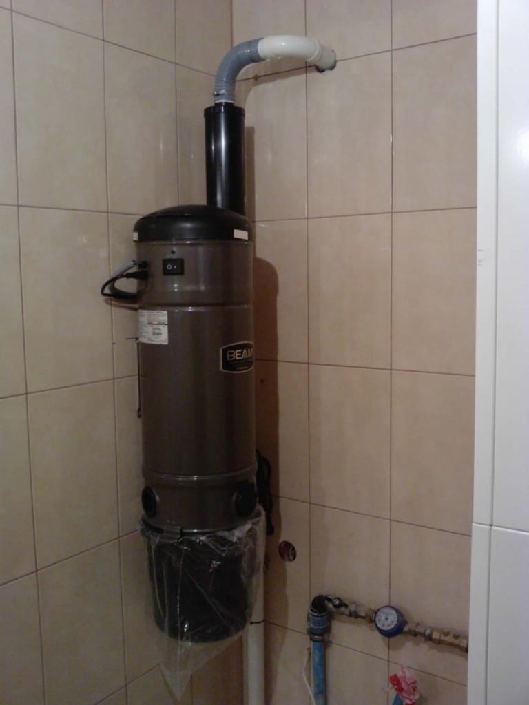 Centralny odkurzacz BEAM zamontowany na kafelkowanej ścianie w łazience, widoczne rury instalacji i worek na kurz. Widok z przodu, lekko z dołu.