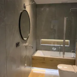 Nowoczesna łazienka z szarymi płytkami, okrągłym lustrem, drewnianą obudową wanny z podświetleniem LED i toaletą myjącą.