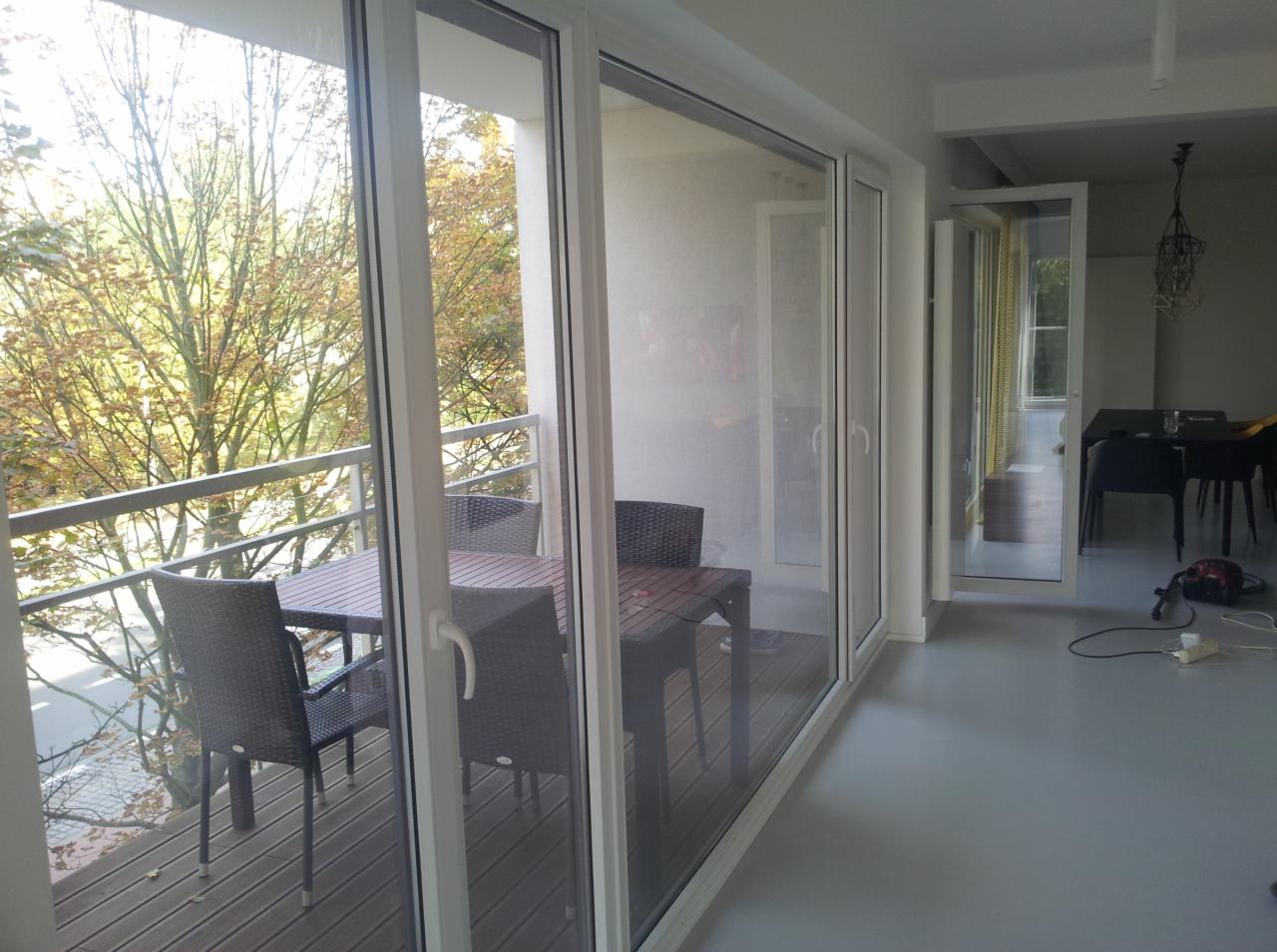 Zadaszony balkon z drewnianą podłogą i meblami ogrodowymi, widoczny przez duże, przesuwne okna. Na zewnątrz drzewa z jesiennymi liśćmi.