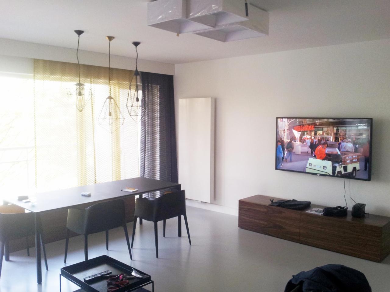 Minimalistyczny salon z szarą podłogą, czarnym stołem i wiszącymi lampami. Telewizor na ścianie, drewniana szafka RTV. Żółto-szare zasłony w oknach.