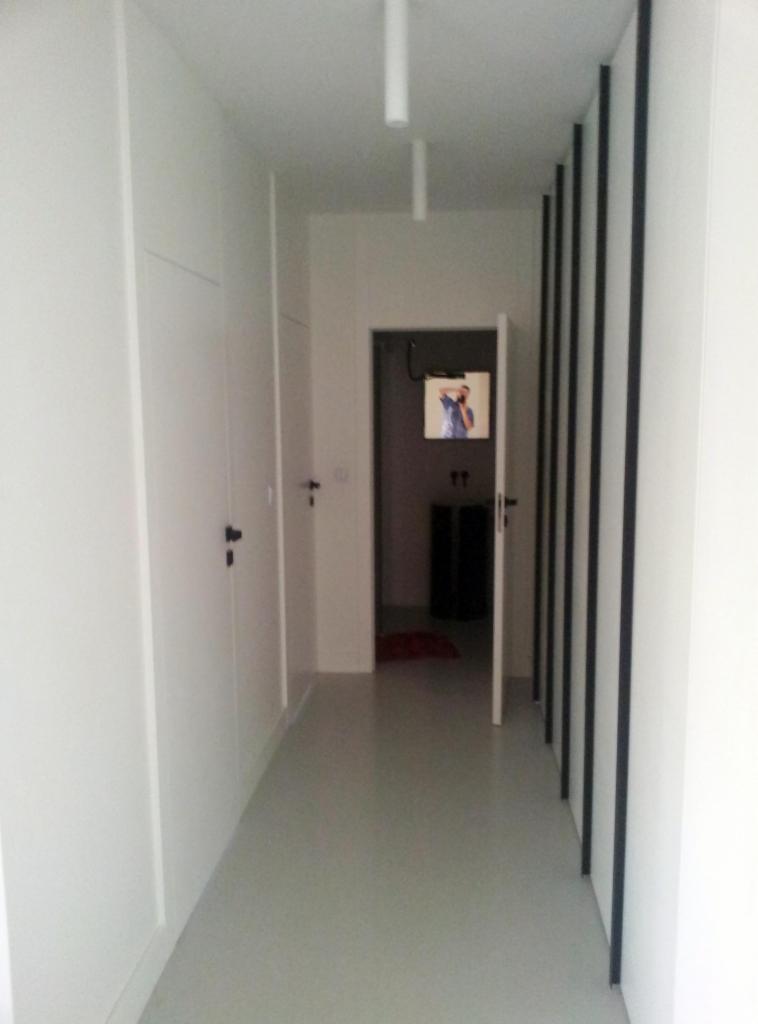 Wnętrze korytarza z jasną posadzką, białymi ścianami i drzwiami z czarnymi klamkami. Widoczny element dekoracyjny z czarnymi pionowymi listwami oraz otwarte drzwi do pokoju.