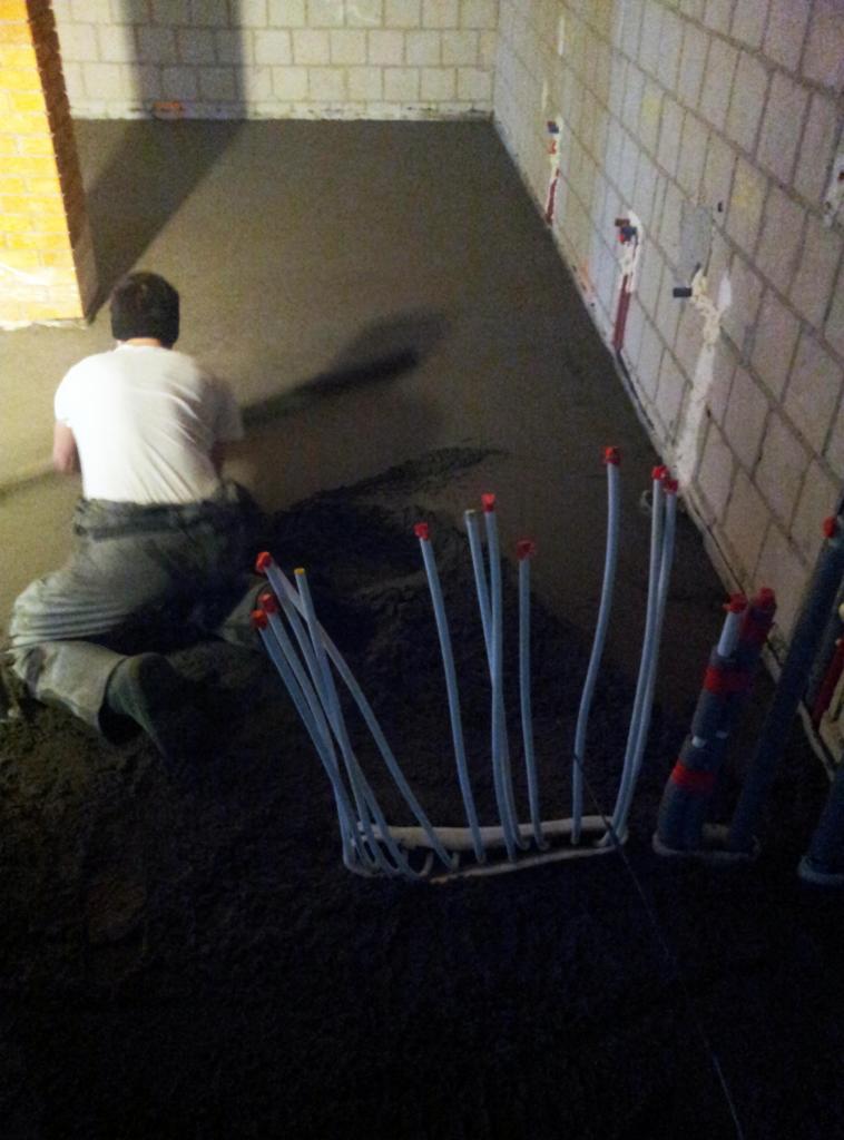 Wyrównywanie wylewki pod posadzkę w pomieszczeniu z instalacją grzewczą podłogową. Widoczne wystające rurki instalacji i pracownik w trakcie rozprowadzania masy.