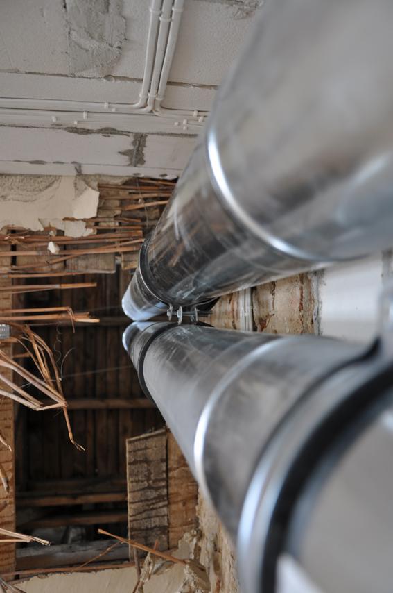 Dwa metalowe przewody wentylacyjne przymocowane do sufitu w trakcie remontu, widoczne instalacje elektryczne i fragmenty starego drewnianego stropu z resztkami tynku.