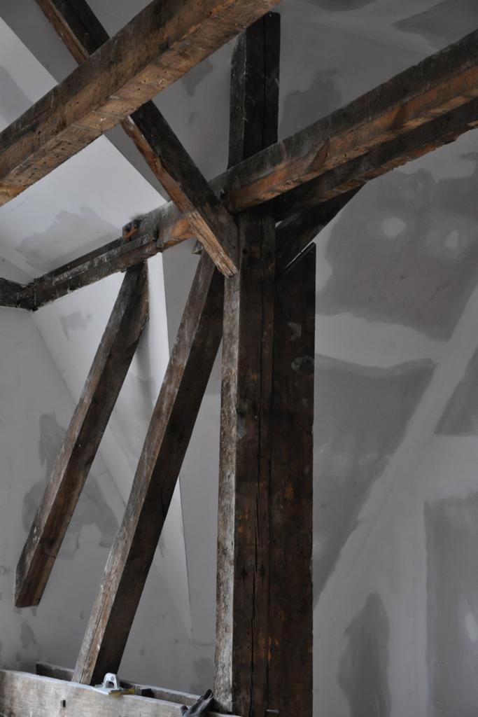 Wnętrze remontowanej kamienicy w Poznaniu: surowe drewniane belki stropowe podtrzymujące konstrukcję dachu, widoczne ślady po starym tynku na ścianach, przygotowywane do dalszych prac wykończeniowych.