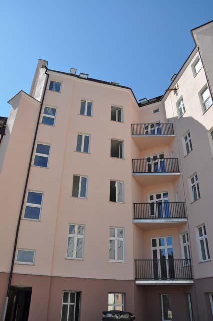 Odnowiona elewacja kamienicy w pastelowych kolorach z ciemnobrązowym cokołem, widok z dołu na balkony z czarnymi balustradami i okna z białymi ramami pod błękitnym niebem.