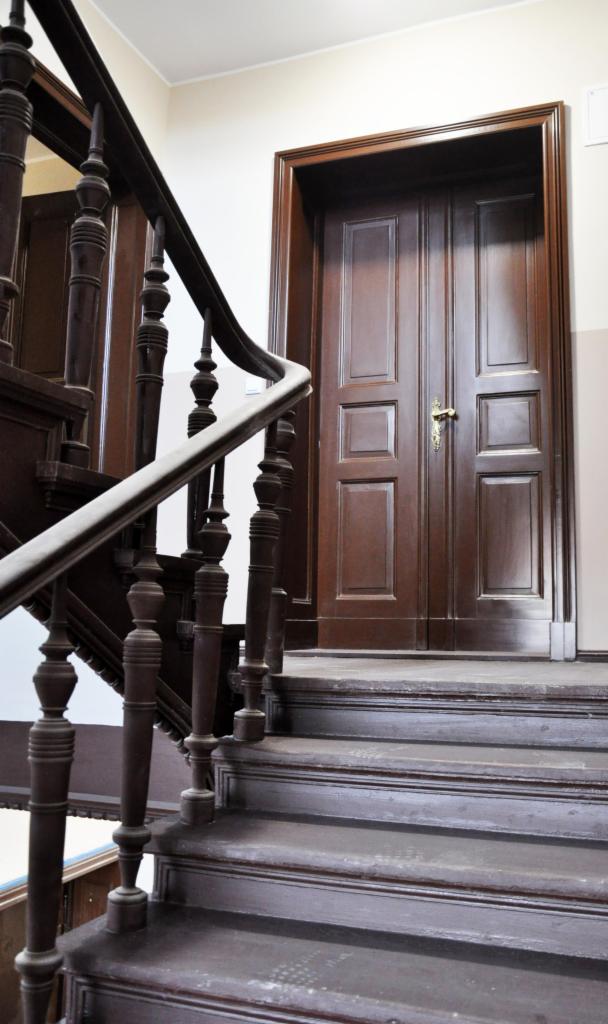 Ciemne, drewniane schody z balustradą prowadzą do dwuskrzydłowych drzwi. Widoczne detale rzeźbione w drewnie i złota klamka. Renowacja starego budownictwa.