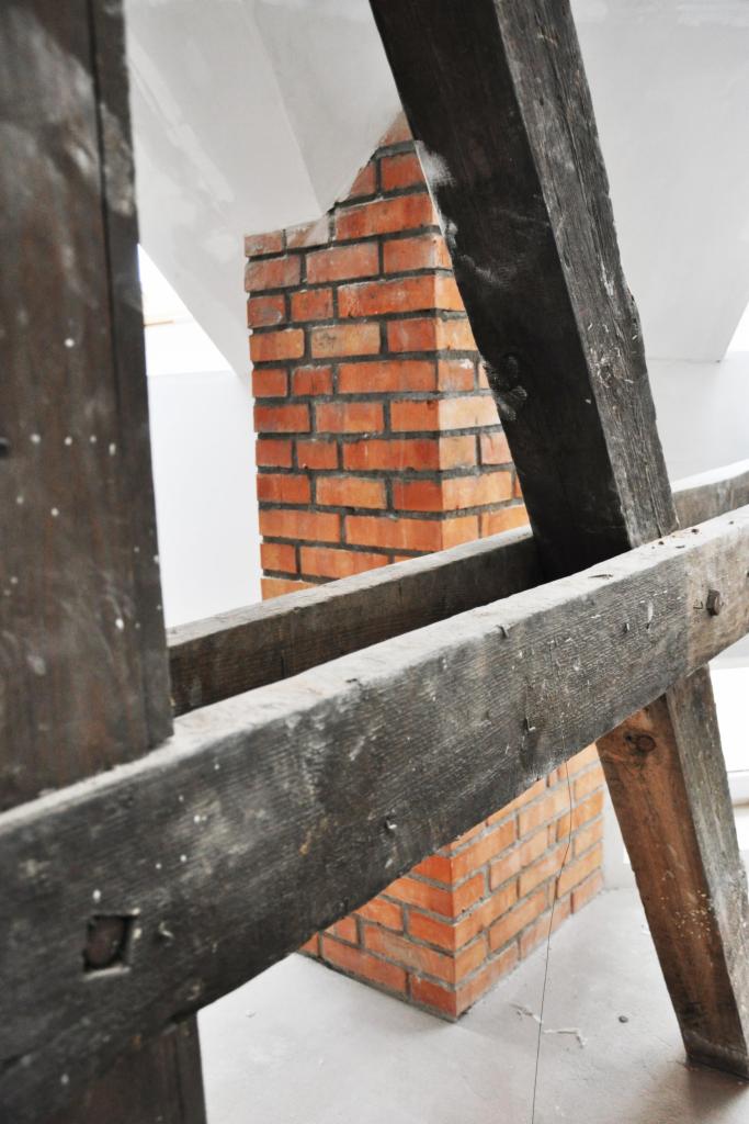 Ceglany komin w surowym wnętrzu poddasza, widoczny przez drewniane elementy konstrukcyjne. Ujęcie z dołu, podkreśla industrialny charakter.