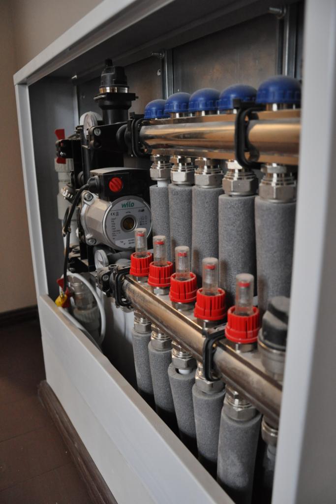 Rozdzielacz ogrzewania podłogowego w szafce z pompą obiegową Wilo, widoczne zawory z niebieskimi i czerwonymi pokrętłami oraz szare rury. Całość w jasnej obudowie.
