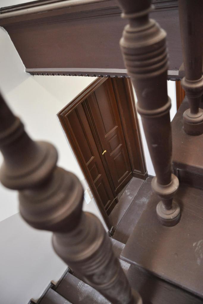 Ujęcie z góry na drewniane schody z balustradą i otwarte drzwi w brązowej ramie. Perspektywa z góry, detale architektoniczne, ciepłe odcienie drewna.