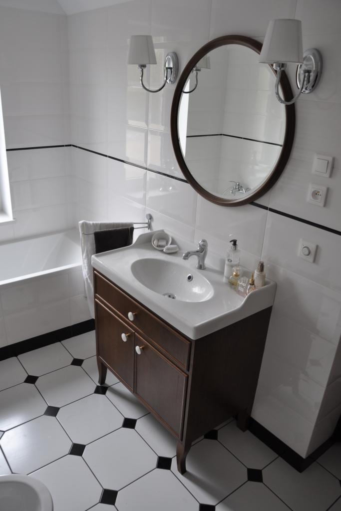 Wykończona łazienka: białe płytki z czarnym akcentem, brązowa szafka pod umywalką z okrągłym lustrem i lampami. Klasyczny styl.