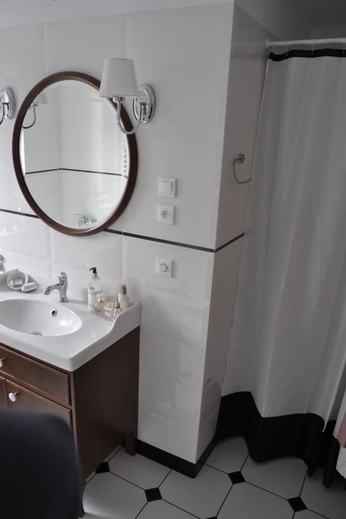 Elegancka łazienka w bieli i brązie: okrągłe lustro z oświetleniem, umywalka z szafką i biało-czarna podłoga. Widoczny fragment zasłony prysznicowej.