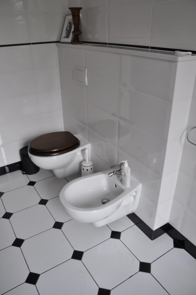 Biała łazienka z toaletą z brązową deską, bidetem, białymi płytkami i czarnymi akcentami na podłodze i ścianach. Na półce dekoracje.