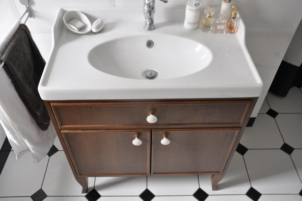 Biała umywalka z brązową szafką w łazience z czarno-białą podłogą. Na umywalce mydło i kosmetyki, obok wiszą ręczniki.