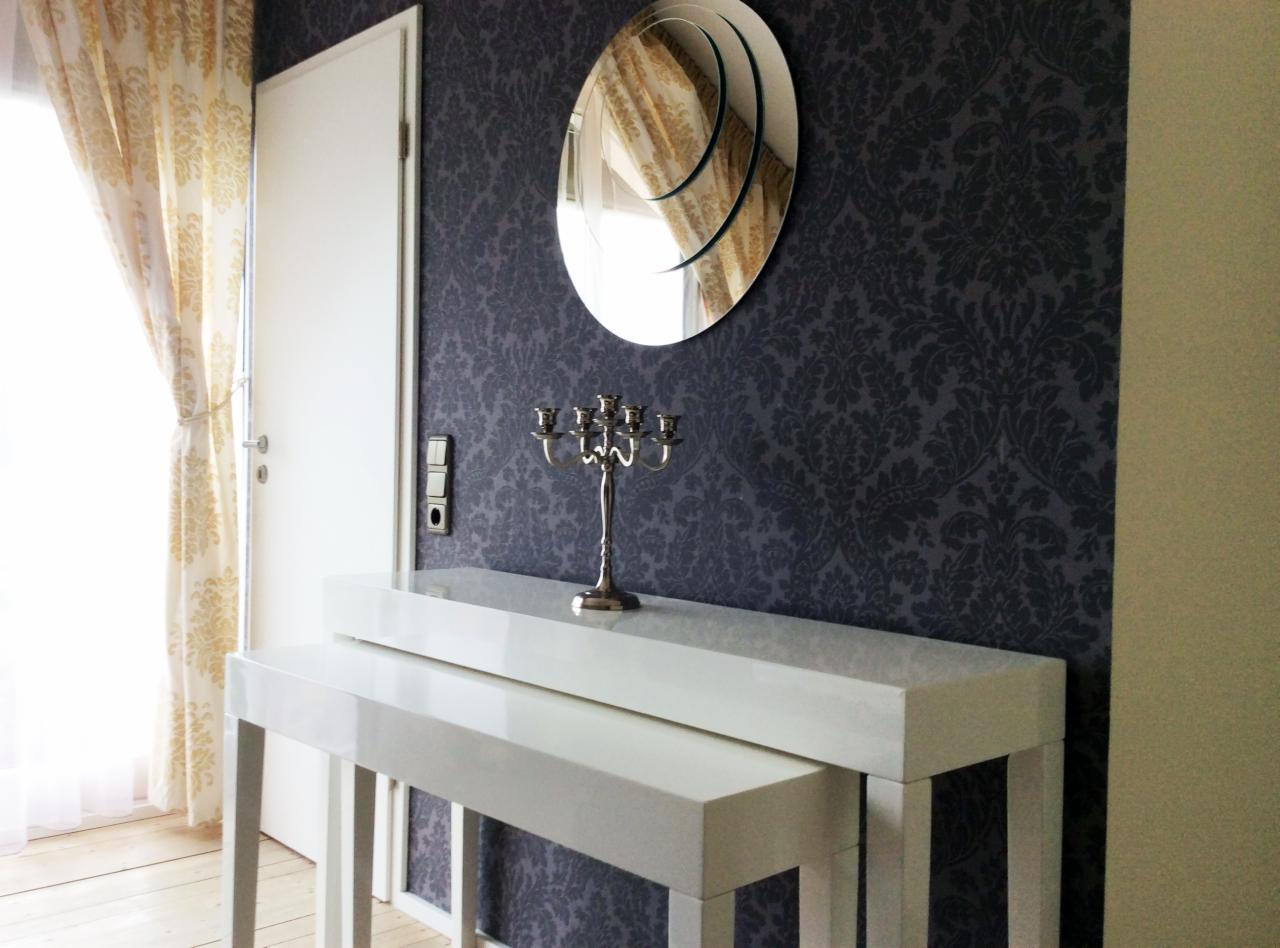 Białe konsole na tle ściany z wzorzystą tapetą, nad nimi okrągłe lustro i srebrny świecznik. Obok drzwi i zasłonięte okno z jasnymi zasłonami.