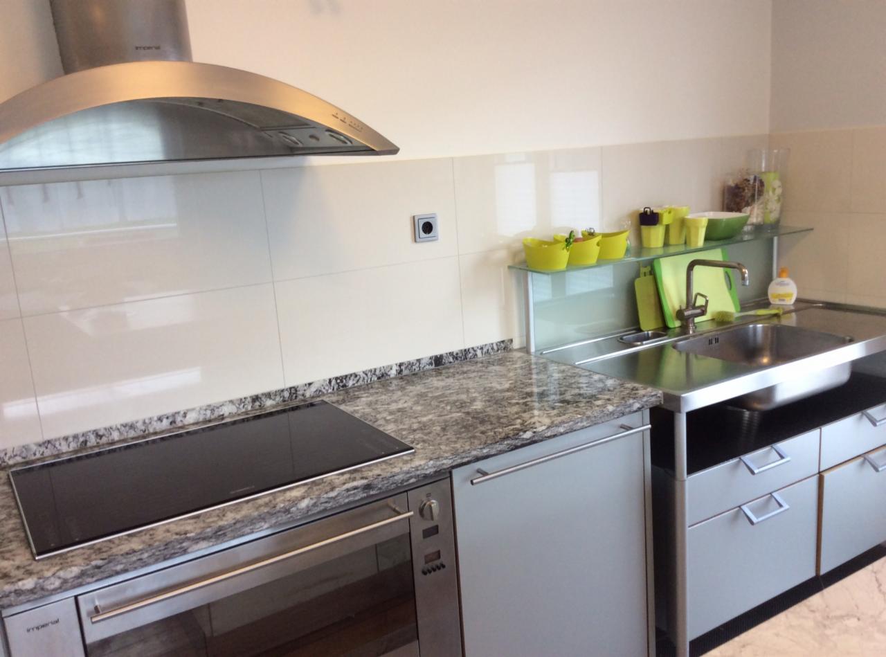 Nowoczesna kuchnia z granitowym blatem, płytą indukcyjną i metalowym okapem. Jasne, kafelkowe ściany i stalowy zlew z baterią. Żółte akcenty kolorystyczne.