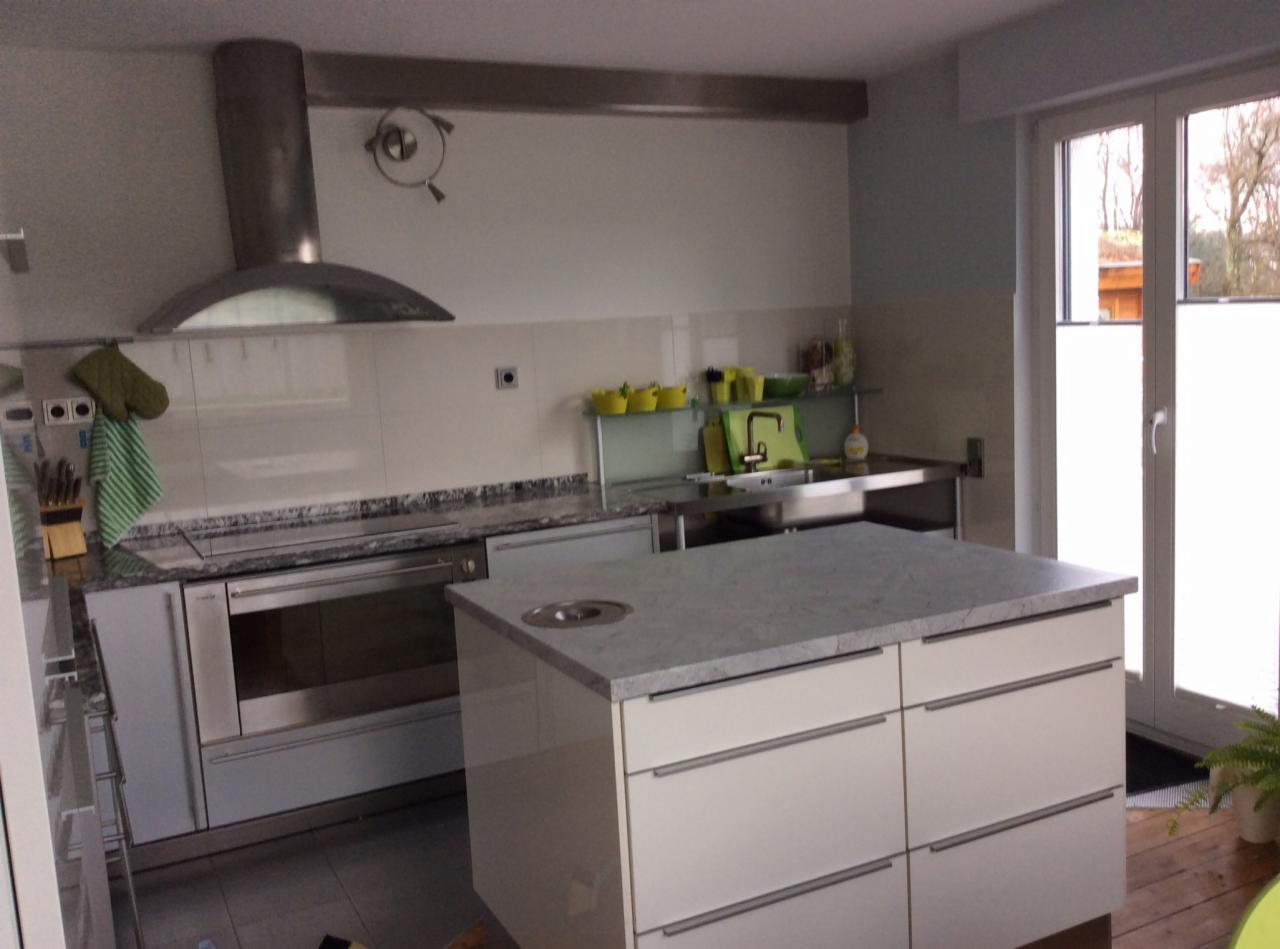 Nowoczesna kuchnia z wyspą i srebrnym okapem. Jasne fronty szafek, granitowy blat, okno z roletą. Minimalistyczny design, elementy ze stali nierdzewnej.