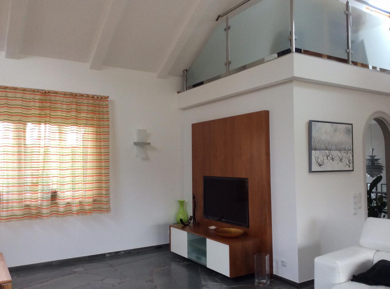 Jasne wnętrze z antresolą i szklaną balustradą. Drewniana zabudowa TV, biała sofa, obraz na ścianie i okno z zasłoną w paski tworzą przytulny charakter.