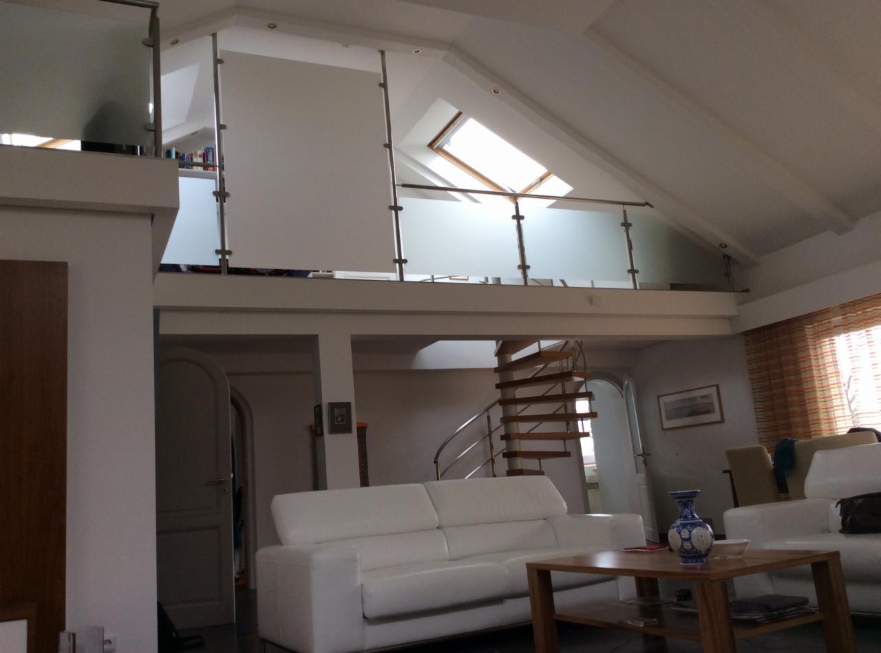 Jasne wnętrze z antresolą zabezpieczoną szklaną balustradą i spiralnymi schodami. Widoczne okno dachowe i białe sofy w części wypoczynkowej.