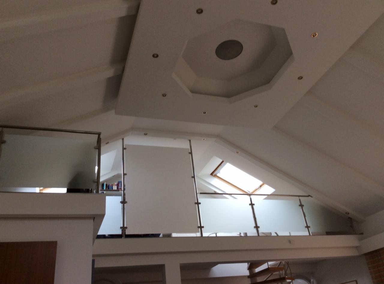 Jasne wnętrze z antresolą i szklaną balustradą, oświetlone światłem z okna dachowego. Sufit zdobiony geometrycznym wzorem z punktowym oświetleniem.