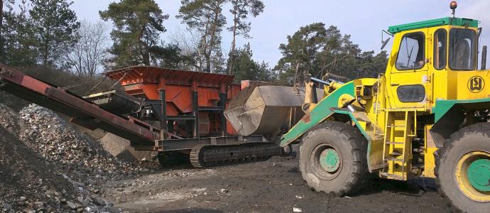 Żółto-zielona ładowarka kołowa z łyżką i pomarańczowa kruszarka na gąsienicach na placu budowy z hałdą gruzu, w tle drzewa.