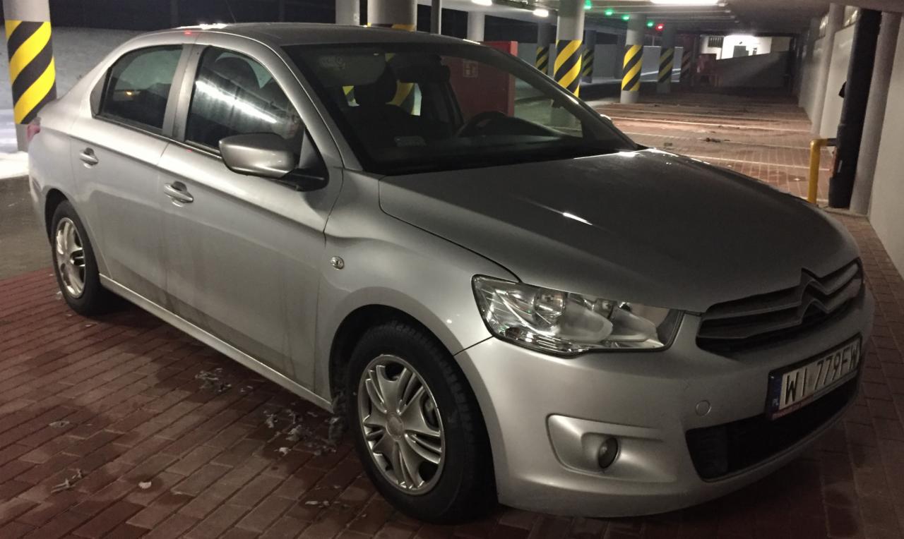 Srebrny sedan na ceglanym parkingu podziemnym, widoczne żółto-czarne słupy i tablica rejestracyjna z Warszawy.