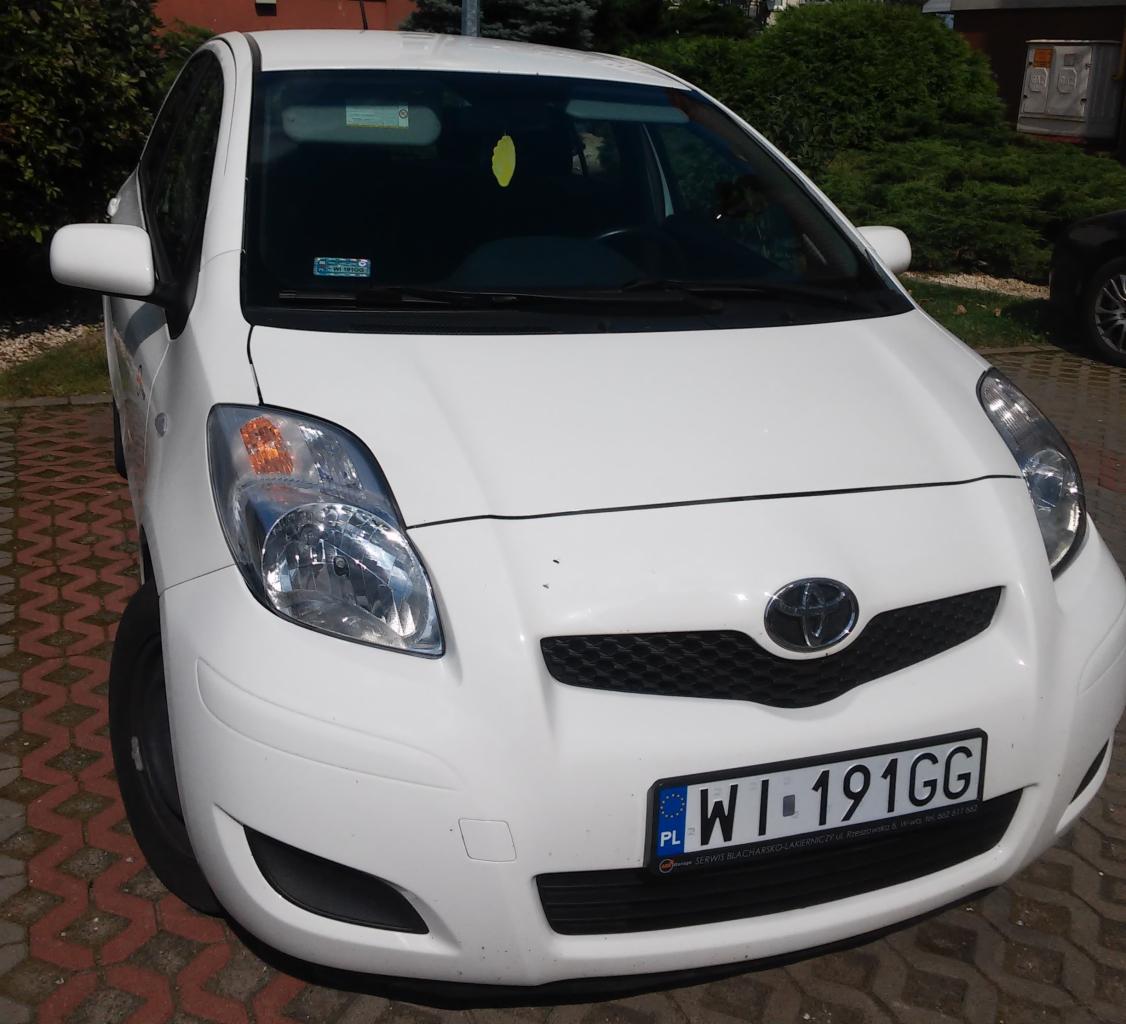Przód białego samochodu Toyota Yaris z widoczną tablicą rejestracyjną na tle brukowanego placu i zieleni.