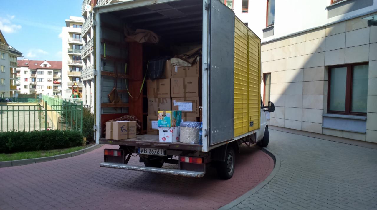 Żółty bus transportowy z otwartą paką, załadowany kartonami, stoi na kostce brukowej przed budynkiem mieszkalnym. Widoczny numer rejestracyjny.