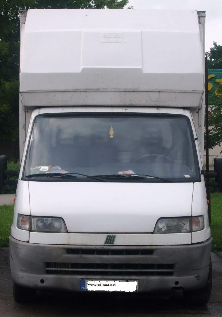 Biały samochód dostawczy Fiat Ducato z nadbudową, widok z przodu, zaparkowany na zewnątrz. Na desce rozdzielczej widoczne dokumenty, a na lusterku wisi zapach.