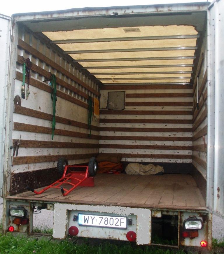 Wnętrze paki busa transportowego z wózkiem transportowym, zabezpieczeniami i tablicą rejestracyjną WY 7802F, widok z zewnątrz.