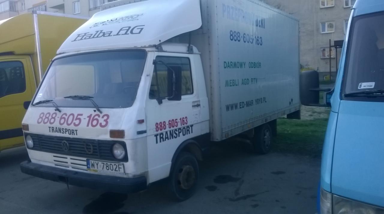 Biały samochód dostawczy z numerem telefonu i adresem strony www, zaparkowany na zewnątrz. Widoczny bok i przód pojazdu z żółtym i niebieskim busem obok.