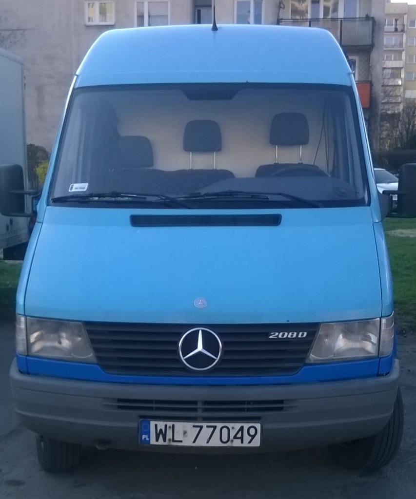 Błękitny Mercedes-Benz 208D z numerem rejestracyjnym WL 77049, zaparkowany na zewnątrz, widok z przodu, budynek w tle.