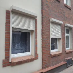 Elewacja budynku z trzema oknami PCV z zamontowanymi zewnętrznymi roletami w kolorze jasnego beżu, z częściowo opuszczoną roletą na pierwszym planie.