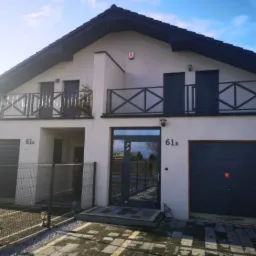 Nowoczesny dom dwurodzinny w Poznaniu z szarymi bramami garażowymi, balkonami z ozdobną balustradą i ciemną stolarką okienną, sfotografowany w słoneczny dzień.