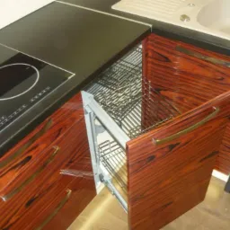 Szafka narożna z wysuwanym koszem i frontem z egzotycznego drewna, wkomponowana w zabudowę kuchenną z czarnym blatem i płytą indukcyjną.