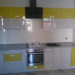 Ż&oacute;łto-szara zabudowa kuchenna z czarnym okapem i płytkami. Fronty szafek z poziomym wzorem. Widoczny piekarnik i zlew.