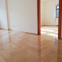 Błyszcząca, jasna podłoga z parkietu ułożonego w jodełkę, widok z niskiej perspektywy, w tle drzwi do kolejnego pomieszczenia z oknem.