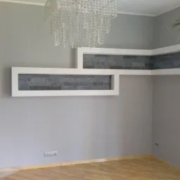 Szara ściana z dekoracyjnymi wnękami wyłożonymi szarą cegłą, kryształowy żyrandol i fragment kominka z kamienia. Na podłodze parkiet.