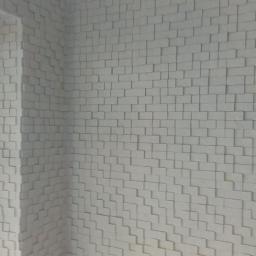 Rab-Bud - Ściana wewnętrzna wykończona trójwymiarowymi, białymi panelami o geometrycznym wzorze, widoczna z perspektywy narożnika pomieszczenia, obok okna.