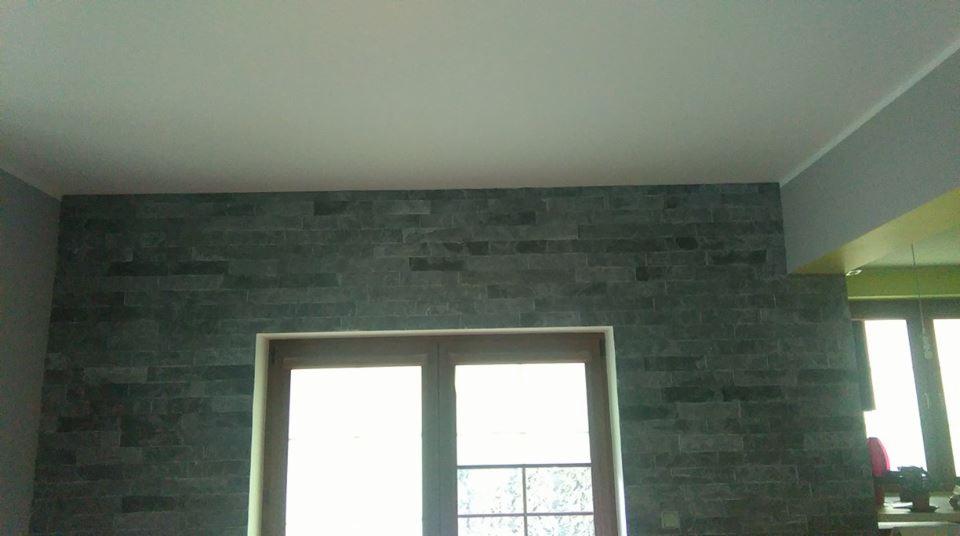 Ściana wykończona szarą, nieregularną cegłą dekoracyjną nad oknem. Widoczne detale faktury i ułożenia cegiełek. Całość w stonowanej kolorystyce.