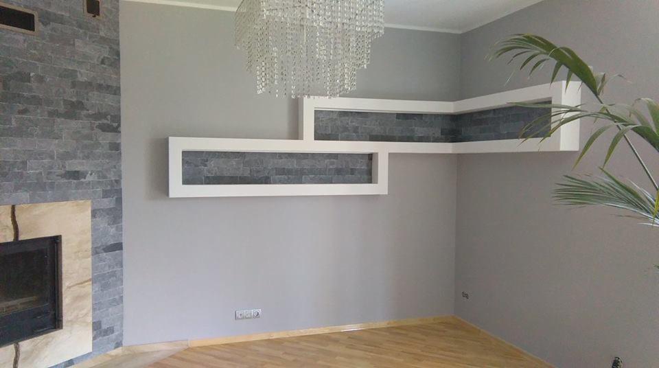 Szara ściana z dekoracyjnymi wnękami wyłożonymi szarą cegłą, kryształowy żyrandol i fragment kominka z kamienia. Na podłodze parkiet.
