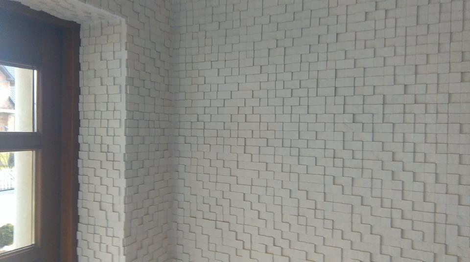 Ściana wewnętrzna wykończona trójwymiarowymi, białymi panelami o geometrycznym wzorze, widoczna z perspektywy narożnika pomieszczenia, obok okna.