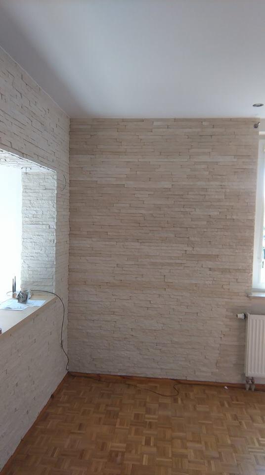 Wnętrze pokoju z wykończoną ścianą dekoracyjną z jasnego kamienia imitującego cegłę. Widoczny fragment parkietu i okno z grzejnikiem.