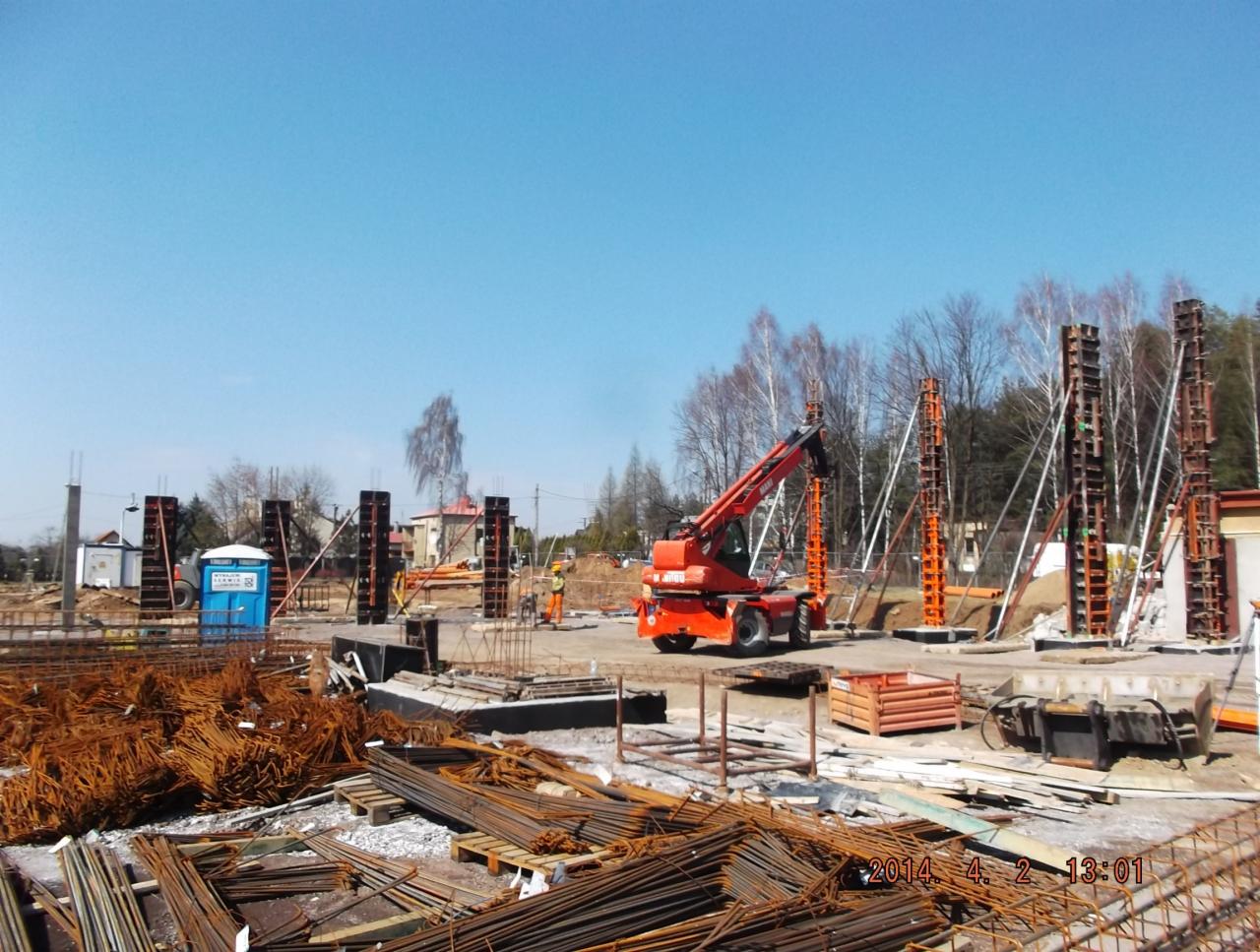 Fundamenty budynku w trakcie wylewania, widoczne zbrojenia, szalunki i dźwig teleskopowy. Na pierwszym planie składowane elementy zbrojeniowe.