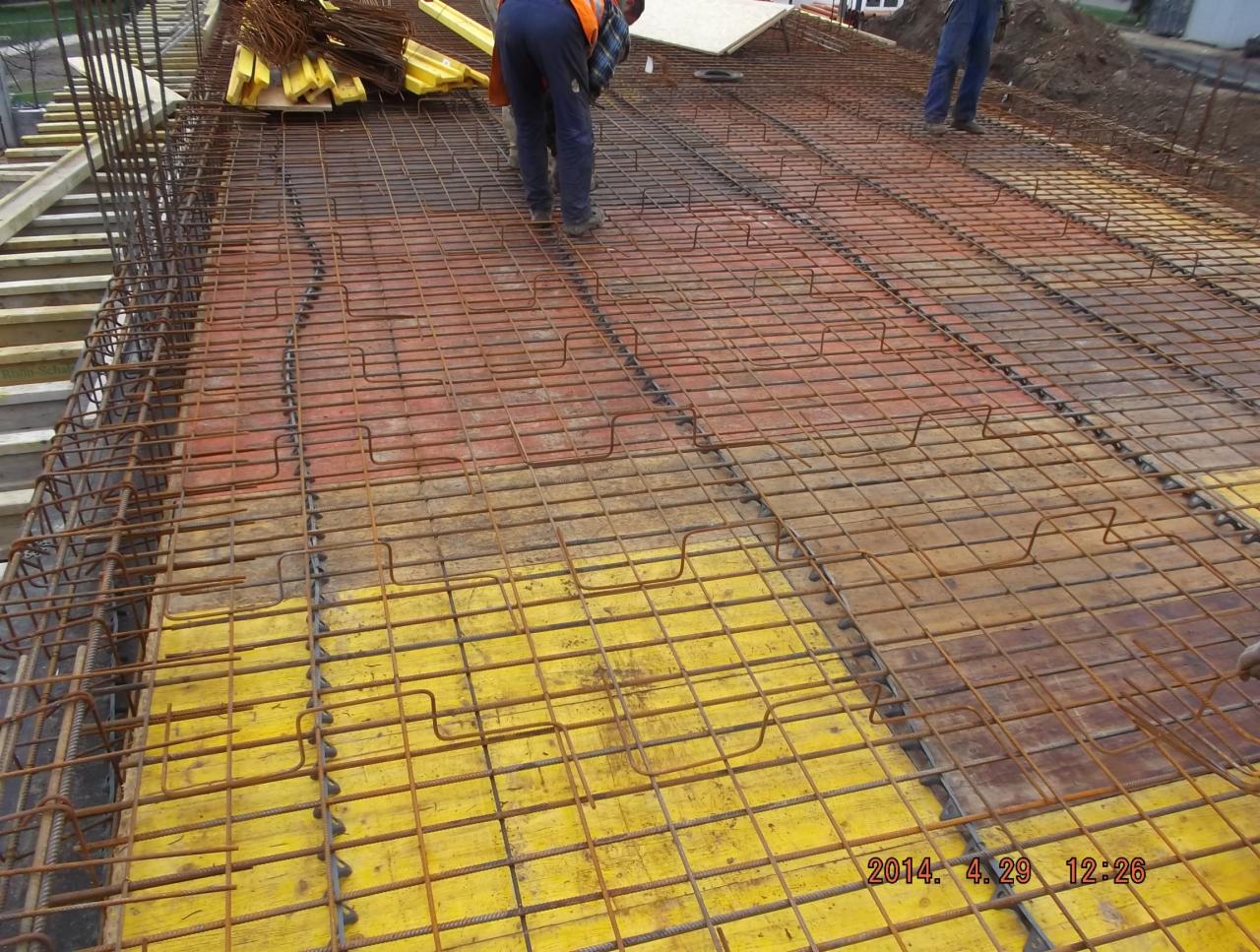 Zbrojenie płyty fundamentowej z widocznymi deskami szalunkowymi w kolorze żółtym i fragmentami ułożonego zbrojenia pionowego. Widoczni pracownicy budowlani.