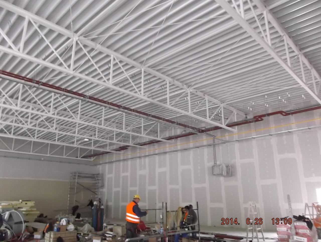 Wnętrze hali w budowie: biała konstrukcja dachu z widocznymi instalacjami, pracownicy w odblaskowych kamizelkach montują ścianę z płyt gipsowo-kartonowych.