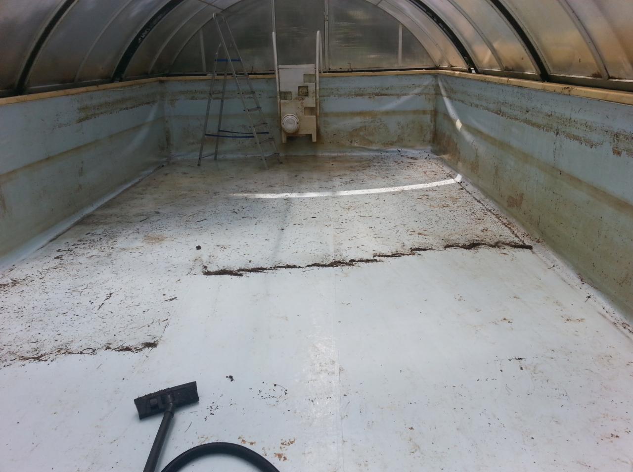 Brudny, pusty basen pod zadaszeniem, z widocznymi zabrudzeniami i resztkami na dnie. Na pierwszym planie fragment odkurzacza. Widoczna drabinka i elementy konstrukcyjne basenu.