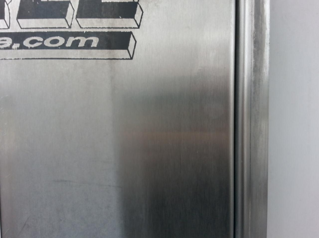 Szczegółowe ujęcie metalowej powierzchni z delikatnym szczotkowaniem i częściowo widocznym logo w lewym górnym rogu, oświetlone naturalnym światłem.